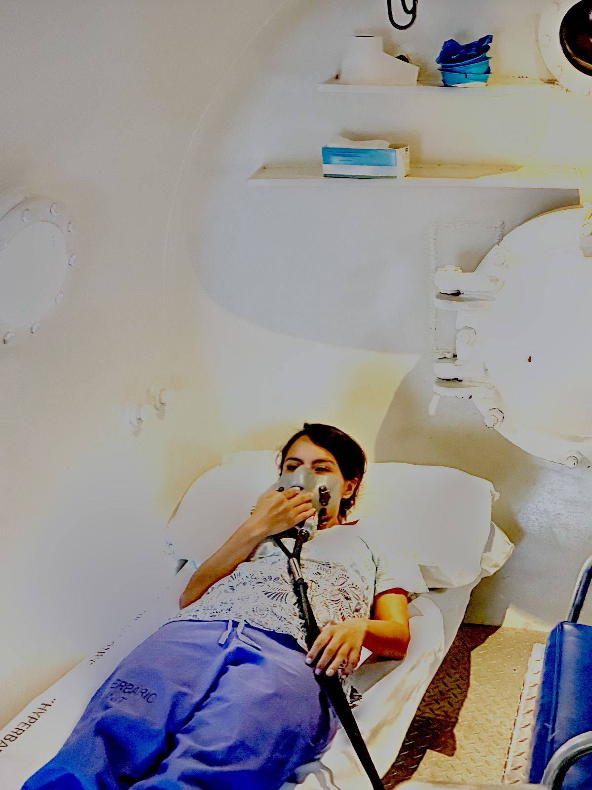 A chamber attendant seated inside a multiplace recompression chamber, holding the hand of a reclined patient